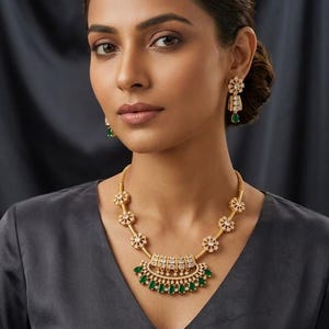 May include: A gold-toned necklace and matching drop earrings set featuring clear white stones and teardrop-shaped green stones. The necklace has a central semi-circular pendant with dangling green stones, connected to floral-shaped links.