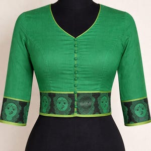 May include: A green blouse with three-quarter sleeves and a V-neck. The blouse features a decorative band with green faces on a black background. The sleeves and neckline are trimmed with a lighter green. The blouse is buttoned down the front.