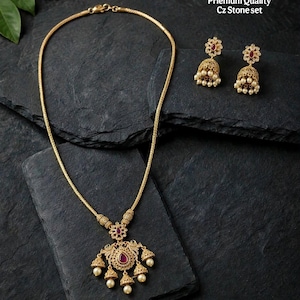 May include: A gold-toned necklace and matching jhumka earring set with a matte finish. The jewelry features red teardrop-shaped stones, clear CZ stones, and small white pearls. Text reads: No.12932-430 Matte Finish Priemum Quality Cz Stone set.