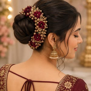 May include: A hair accessory featuring deep burgundy fabric flowers interspersed with clusters of small metallic gold beads. The piece is arranged in a curved shape and pinned into a dark brown hair bun. A gold jhumka earring with small white pearls hangs from the ear.