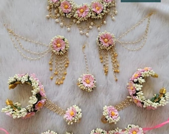 Könnte beinhalten: Ein floraler Schmuck-Set mit Halskette, Ohrringen, Kopfschmuck, Armbändern und Ringen. Das Set umfasst rosa und weiße Blumen mit gelben Zentren, akzentuiert mit Gold- und Perlendetails. Der Schmuck ist auf einer weißen, strukturierten Oberfläche angeordnet.