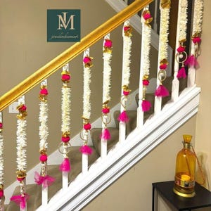 Puede incluir: Escalera decorada con guirnaldas blancas, detalles dorados y borlas rosas vibrantes. Las guirnaldas cuelgan de los balaustres blancos y el pasamanos dorado. Una linterna decorativa está sobre una mesa auxiliar negra. El logotipo "M jewelindiamart" es visible.