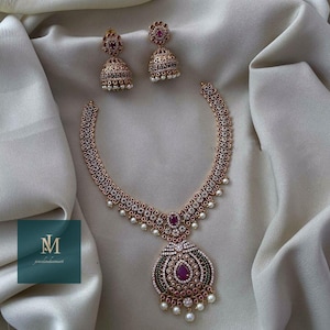 May include: A gold-coloured jewellery set comprising a necklace and matching earrings. The necklace features a large pendant with a central ruby-coloured stone, surrounded by smaller stones and pearls. The earrings are bell-shaped with similar detailing. The set is displayed on white fabric.