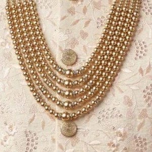 May include: A multi-strand gold-toned beaded necklace with a layered design. The necklace is worn over a cream-colored embroidered garment with gold buttons. The image includes a logo that reads "jewelindiamart".