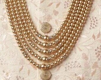 May include: A multi-strand gold-toned beaded necklace with a layered design. The necklace is worn over a cream-colored embroidered garment with gold buttons. The image includes a logo that reads "jewelindiamart".