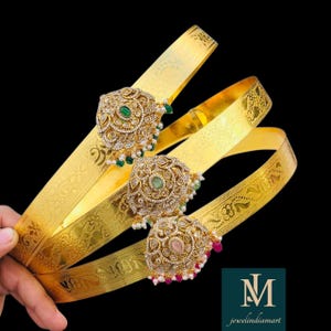 May include: Three gold waist belts with elaborate, jewel-adorned centrepieces. Each centrepiece has a central gemstone, surrounded by smaller gems and pearls. The belts have a textured design and are set against a black background.