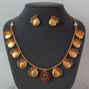 May include: A brass necklace and earring set with a matte finish. The necklace features a central floral pendant with red and green stones, flanked by teardrop-shaped charms with green and red accents. Text reads: No.1102-300, Matte Finish, Priemum Quality, Made In Brass.