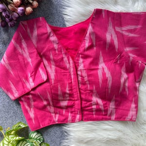 May include: A vibrant pink blouse with a V-neck and short sleeves. The fabric features a white ikat pattern. The blouse is unbuttoned, revealing a deeper pink lining. The garment is displayed on a white fur-like surface.