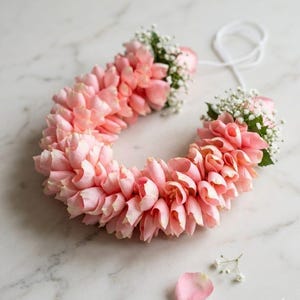 May include: A U-shaped garland made of pink rose petals and small white flowers. The floral arrangement has a white string attached. The petals are a soft pink colour, and the small white flowers add a delicate touch. A single petal and small flowers are scattered on the surface.