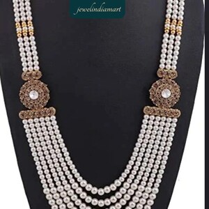 May include: A multi-strand pearl necklace with gold-toned accents and a central decorative element featuring a clear stone. The necklace is displayed on a black mannequin.