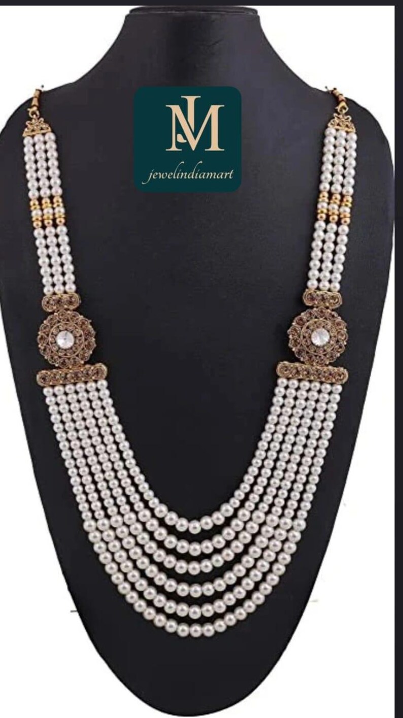 May include: A multi-strand pearl necklace with gold-toned accents and a central decorative element featuring a clear stone. The necklace is displayed on a black mannequin.