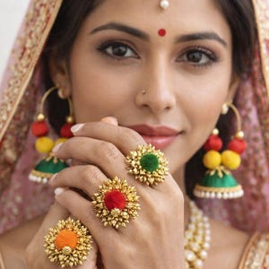 Op de afbeelding: Een vrouw draagt gouden sieraden, waaronder oorbellen met rode, gele en groene accenten, een hoofddeksel en ringen met kleurrijke middelpuntstukken. De ringen hebben oranje, rode en groene accenten. De vrouw draagt een roze en gouden hoofdbedekking.