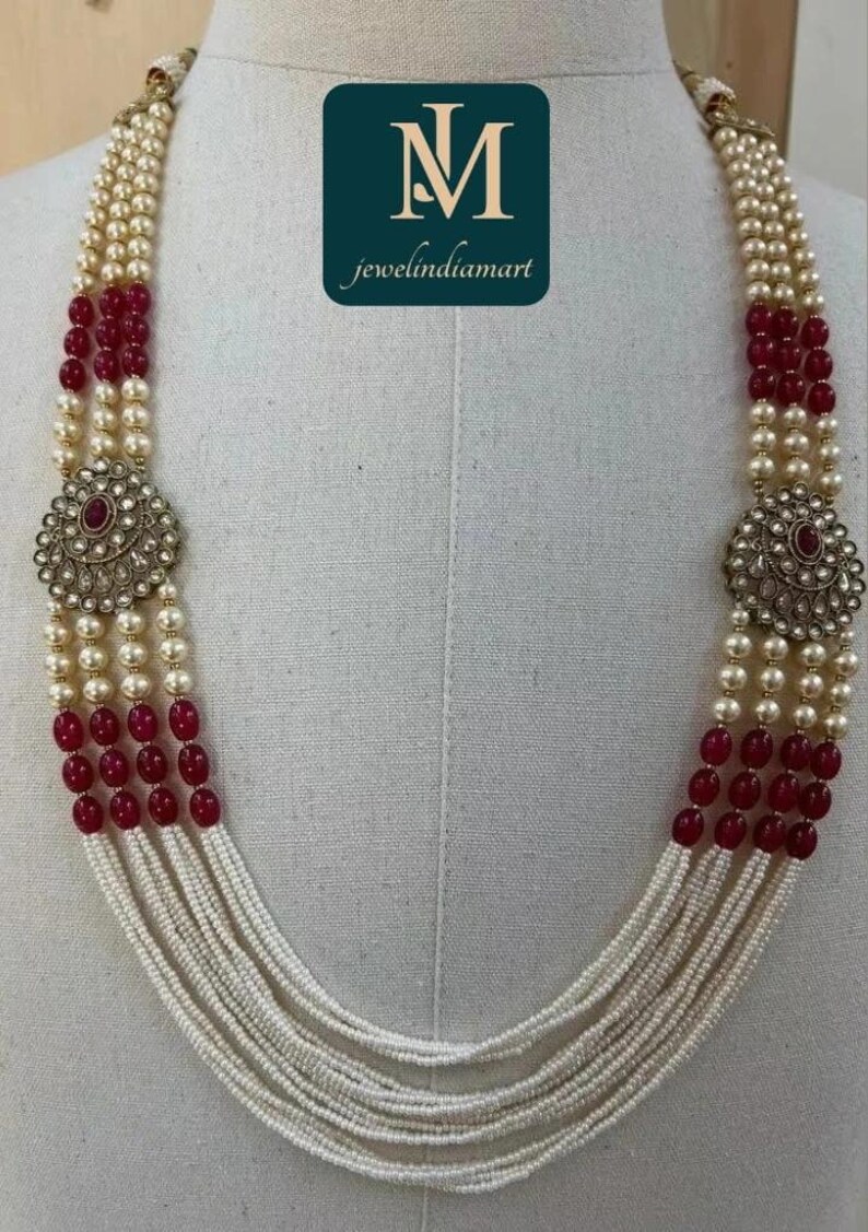 May include: A multi-strand necklace featuring cream-colored pearls, interspersed with red beads. The necklace includes two ornate, circular pendants with a central red stone. The logo "jewelindiamart" is visible.