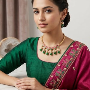 Puede incluir: Una mujer con una blusa verde y un sari magenta con bordados florales. Está adornada con un collar de oro y pendientes a juego con piedras preciosas verdes y rojas y perlas blancas. Su cabello está peinado en un moño trenzado.