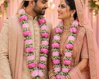 May include: A couple in traditional wedding attire. The individual on the left wears a beige sherwani with gold embroidery and a matching turban. The individual on the right wears a pink lehenga with gold embroidery and a veil. Both wear floral garlands.