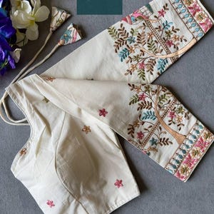 May include: Cream Khadi cotton blouse with detailed embroidery. The long sleeves showcase a tree design with colourful floral and bird accents. The blouse has a round neckline and delicate floral embroidery. The text "Khadi Cotton Blouse" is visible.