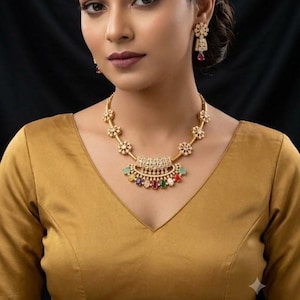 May include: A gold-toned necklace featuring a central semi-circular pendant adorned with clear stones and a row of multi-colored oval gemstones in red, green, and white. The chain includes floral-shaped links. Matching drop earrings are included.