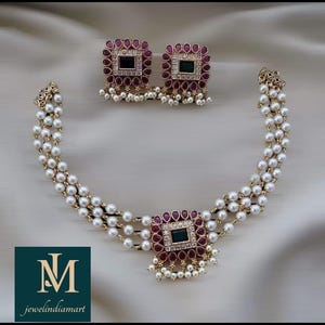 May include: A gold-coloured necklace and earring set. The necklace has three strands of white pearls and a square pendant with a dark green centre stone, surrounded by red and clear stones. The earrings match the pendant design.