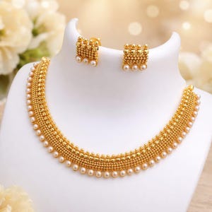 May include: A gold-colored necklace and matching earrings set displayed on a white bust. The necklace and earrings feature a beaded design with small, round pearls. The jewelry is presented against a soft, blurred background.