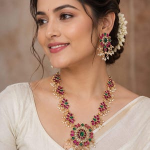 May include: A gold-toned necklace and matching earring set featuring floral-shaped motifs inlaid with pink and green stones. The jewelry pieces are accented with small white beads hanging from the edges, creating a detailed, layered appearance.