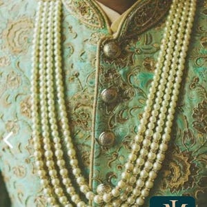 May include: A multi-strand pearl necklace with gold accents adorns a teal and gold embroidered jacket. The jacket features ornate gold detailing and large, decorative buttons. The image includes the text "jewelindiamart".