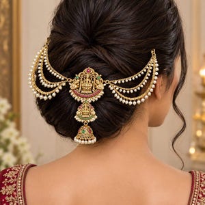 Könnte beinhalten: Ein goldfarbenes Haaraccessoire mit einem zentralen Anhänger, der eine Gottheit darstellt und mit roten und grünen Steinen verziert ist. Es umfasst mehrere drapierte Ketten mit kleinen weißen Perlen und einen glockenförmigen Anhänger.