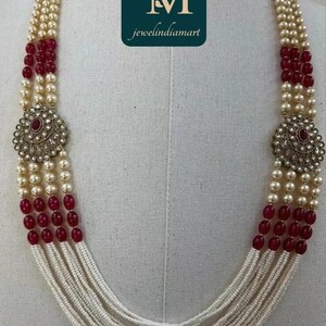 May include: A multi-strand necklace featuring cream-colored pearls, interspersed with red beads. The necklace includes two ornate, circular pendants with a central red stone. The logo "jewelindiamart" is visible.
