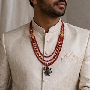 May include: A layered necklace with red beads and gold details. The necklace has a pendant with a figure. The necklace is worn over a cream jacket.