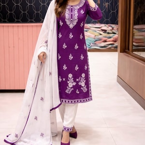 May include: A purple embroidered tunic and white pants ensemble with a matching white scarf. The tunic features intricate white floral embroidery around the neckline, sleeves, and bodice. The outfit is paired with purple pointed-toe heels.