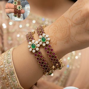 May include: Gold-toned bangles with ruby and emerald accents. The bangles feature a floral design with emerald centers and are adorned with small rubies. The image also shows a smaller set of bangles and the text "jewelindiamart".