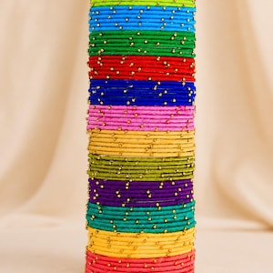 May include: A stack of colorful bangles in various shades, including green, blue, red, pink, and yellow. Each bangle is adorned with small, gold-colored beads. The bangles are stacked vertically, creating a vibrant and eye-catching display.