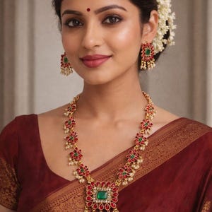 May include: A woman wearing a gold-toned necklace and earrings set with red and green floral designs and a central green square stone. The jewelry complements a maroon sari with gold trim. The image includes text: No905 230 Matte Finish Premium Quality.