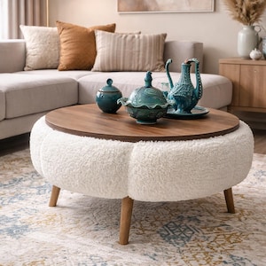 May include: A round ottoman with a white, textured exterior and a dark wood top. The ottoman has wooden legs and is surrounded by decorative items, including teal ceramic pieces and pillows. The room has a neutral color palette.