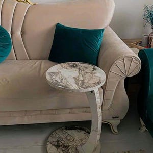 May include: A marble-topped side table with a curved, cream-colored base. The table has a round top and a matching round base, connected by a curved support. The table is placed next to a beige sofa with a teal velvet pillow.