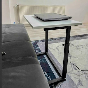 May include: A white and black adjustable laptop table with a black metal frame. A gray laptop rests on the table's surface. The table is positioned next to a gray sofa. The table's height is adjustable.