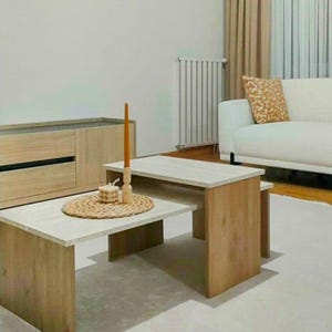 May include: Two-piece wooden coffee table set with a light-colored tabletop and natural wood legs. A woven placemat with a candle and decorative items sits on the lower table. The room features a white sofa and neutral decor.