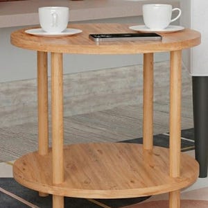 May include: A light brown, two-tiered round side table. The top tier holds two white teacups on saucers and a smartphone. The table has four cylindrical legs and a lower shelf. The table is made of wood and sits on a patterned rug.