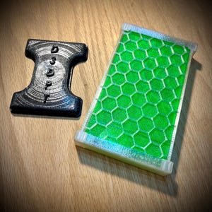 May include: Two 3D-printed items on a wooden surface. One is a black, I-shaped object with the word "DADDY" printed on it. The other is a rectangular, clear object with a green honeycomb pattern.