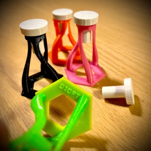 Op de afbeelding: Een verzameling 3D-geprinte objecten in verschillende kleuren, waaronder zwart, oranje en roze. Elk object heeft een cilindrische bovenkant en een uniek basisontwerp. Een losstaand wit cilindrisch stuk en een groen zeshoekig stuk met de tekst "D3DPT" zijn ook aanwezig.