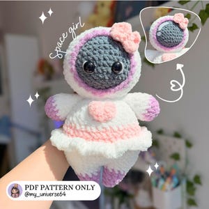 May include: A handmade crochet doll in the shape of a space girl. The doll features a gray head with large black eyes, a white spacesuit with pink accents, and a pink bow. The text "space girl" is written on the image.