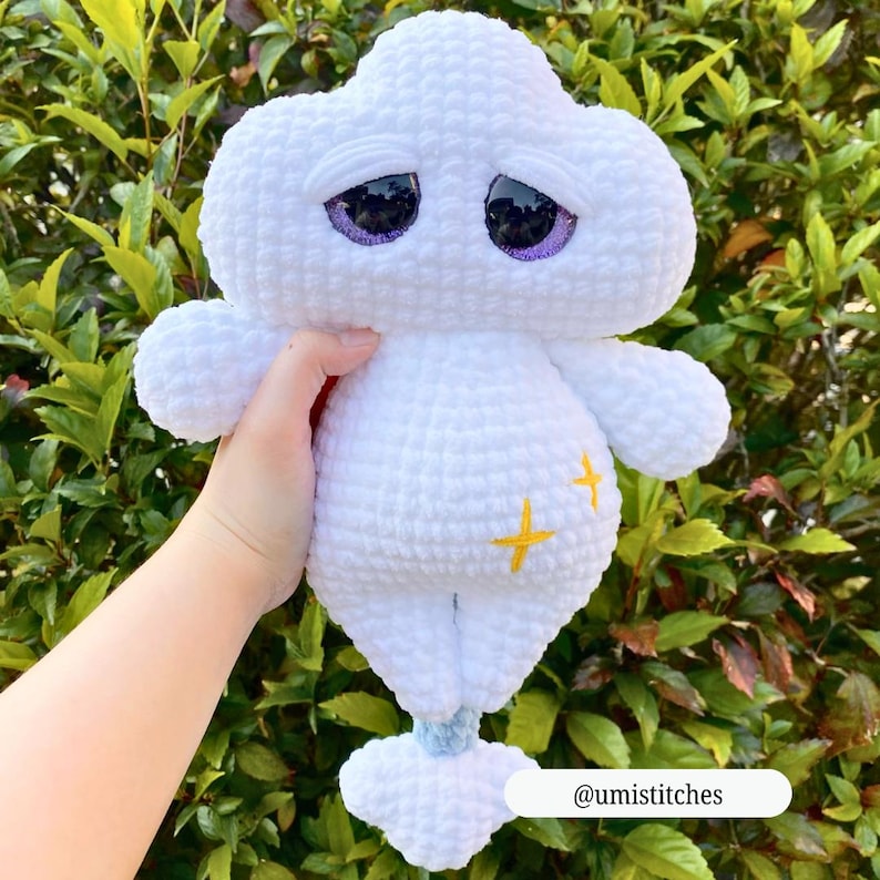 Op de afbeelding: Een handgemaakte witte gehaakte wolk knuffel met paarse ogen en gele kruis accenten. Het wolkvormige speelgoed heeft een verdrietige uitdrukking en een klein blauw detail aan de onderkant. De knuffel wordt vastgehouden tegen een achtergrond van groen gebladerte.