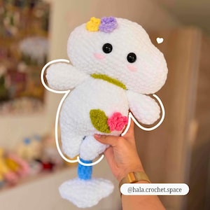 Op de afbeelding: Een handgemaakt wit gehaakt pluche speelgoed met zwarte ogen, roze wangen en kleurrijke bloemenaccenten. Het speelgoed heeft een grillig ontwerp met een blauw-witte basis. De Instagram-handle @hala.crochet.space is zichtbaar.