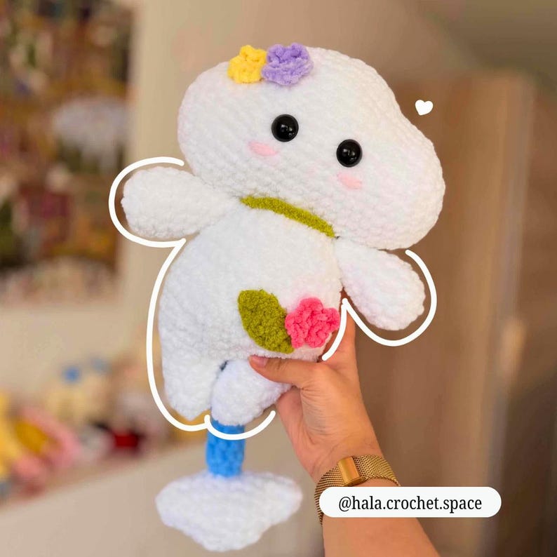 Op de afbeelding: Een handgemaakt wit gehaakt pluche speelgoed met zwarte ogen, roze wangen en kleurrijke bloemenaccenten. Het speelgoed heeft een grillig ontwerp met een blauw-witte basis. De Instagram-handle @hala.crochet.space is zichtbaar.