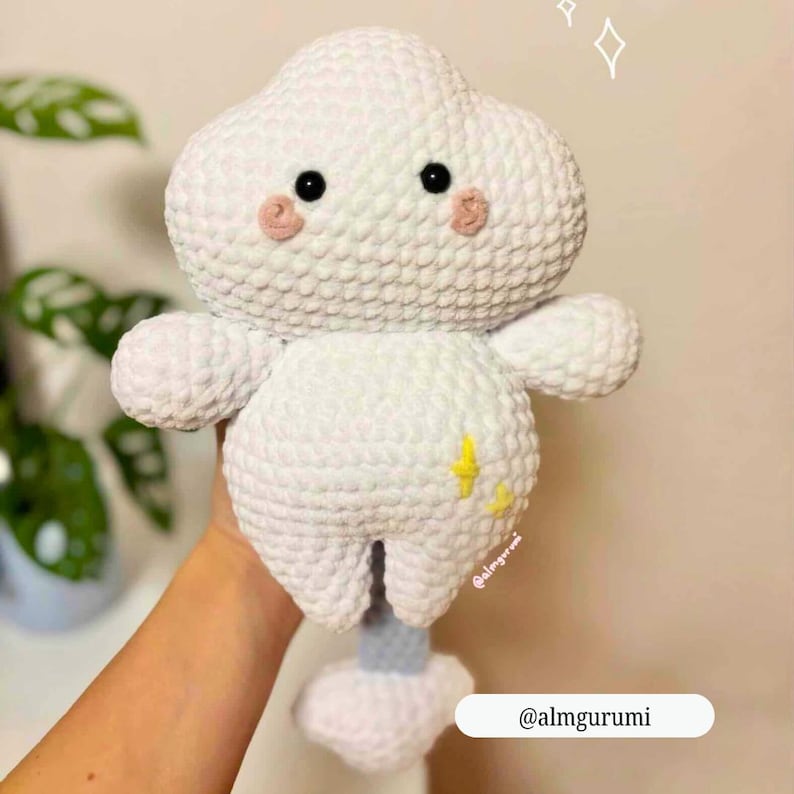 Op de afbeelding: Een handgemaakte witte gehaakte wolk knuffel met zwarte ogen en roze wangen. De wolk heeft kleine armen en benen en een gele bliksem ontwerp. De knuffel wordt vastgehouden. De tekst "@almgurumi" is zichtbaar.