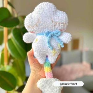 Op de afbeelding: Een handgemaakt wit gehaakt speelgoed in de vorm van een wolk met een blauw lint en een regenboogkleurige onderkant. De wolk heeft geborduurde details en kleine armen. Het speelgoed heeft de tekst "@lolivicrochet" op de bodem.