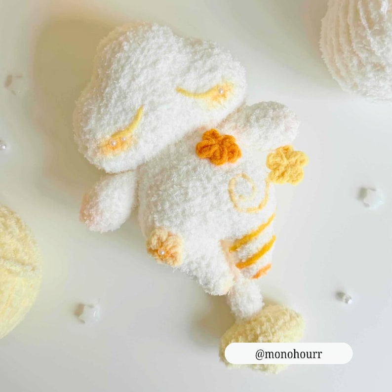 Op de afbeelding: Een handgemaakt wit pluche speelgoed in de vorm van een wolk met oranje en gele accenten. Het speelgoed heeft geborduurde details en decoratieve bloemvormige versieringen. De tekst "@monohourr" is rechtsonder zichtbaar.