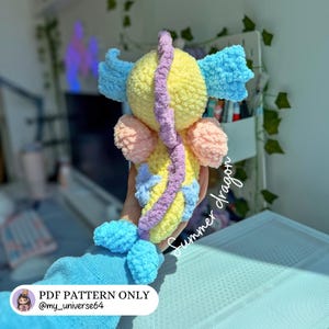 Op de afbeelding: Een handgemaakte gehaakte draak met een geel lichaam, lichtblauwe vinnen en staart, roze zijvleugels en een paarse rugkam. De tekst Summer dragon staat in wit cursief schrift en een label vermeldt PDF PATTERN ONLY @my_universe64.