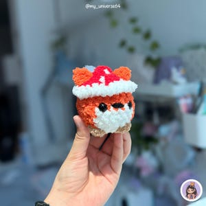 May include: A small, round, crocheted plush toy shaped like a fox with orange, white, and red yarn. The fox features a white chest, black bead eyes, and a red and white hat-like top. The texture is fuzzy and soft, crafted from yarn.