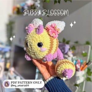 May include: A crocheted bee toy in yellow and brown stripes, with pink and white details. The text "BUZZY BLOSSOM" is above the bee. The bottom of the image reads "PDF PATTERN ONLY @my_universe64".