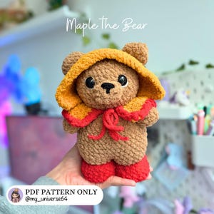 May include: A crocheted brown bear plush toy named Maple The Bear, featuring a yellow hood with a red trim and matching red boots. The bear has large black eyes and a stitched nose. The image includes text reading Maple The Bear and PDF PATTERN ONLY.